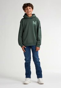 Sudadera con capucha verde con un gráfico frontal y cordón, combinada con jeans azules y zapatillas blancas. La sudadera tiene puños y dobladillo acanalados.