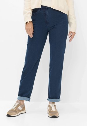 Person iført mørkeblå bukser med opkrogede ben, beige og hvide sneakers, cremefarvet sweater og guldbøjler stående mod en hvid baggrund.