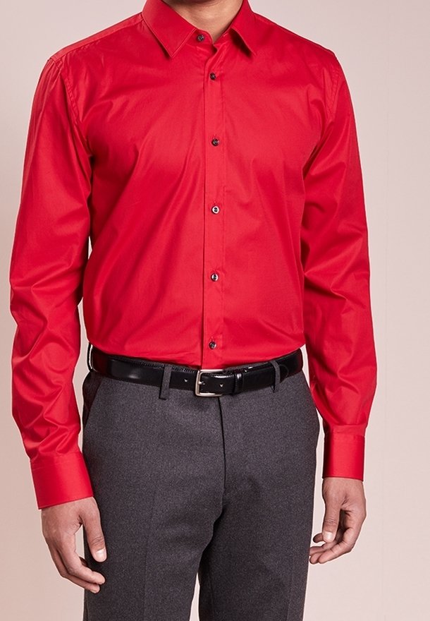 Homme portant une chemise de dress rouge vif à boutons, rentrée dans un pantalon gris avec une ceinture en cuir noir, se tenant devant un fond uni.