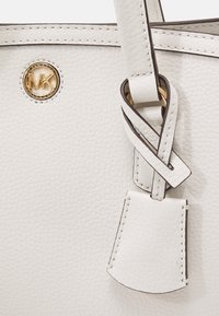 Biała torebka skórzana o teksturowanej powierzchni z logo MK w kolorze złotym, dekoracyjnym węzłem na pasku oraz jasnobrązowym akcentem w szwach.