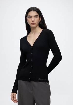 Femme aux longs cheveux foncés portant un cardigan noir boutonné à col en V et un pantalon gris, debout devant un fond clair uni.