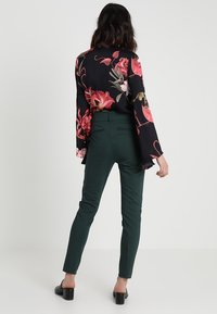 Svart blus med blommönster och trumpetärmar, matchad med gröna slim-fit-byxor och svarta klackskor, sedd bakifrån.