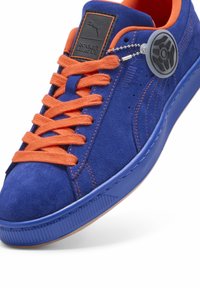 Niebieskie zamszowe sneakersy z pomarańczowymi sznurówkami, gumową podeszwą i czarną metką. Posiadają okrągły emblemat przymocowany łańcuchem do sznurowania.