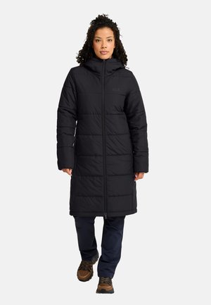 Donna che cammina in avanti indossando un lungo cappotto imbottito nero con cappuccio, pantaloni blu navy e scarpe da trekking marroni su uno sfondo chiaro e uniforme.