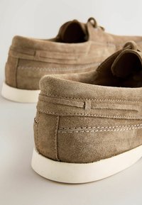Paire de mocassins en daim marron clair avec semelles en caoutchouc blanc vus de dos sur une surface claire.