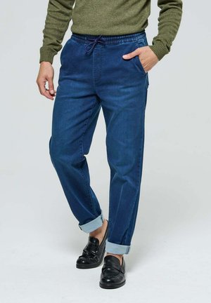 Joggers en denim bleu foncé avec une taille élastique, un cordon de serrage, des poches latérales et des bas retroussés, associés à des mocassins noirs.