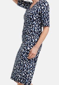 Marinblå klänning med korta ärmar, med ett geometriskt mönster i ljusblått och vitt. Tyget är slätt; designen inkluderar sidoruching.