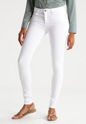 Persoon die witte skinny jeans draagt, een saliegroene longsleeve en bruine platte sandalen met bandjes, staand tegen een witte achtergrond.