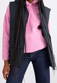 Vrouw in een roze fleecetrui, donkerblauwe jeans en een mouwloze zwarte gewatteerde vest met lange gevlochten haren over haar schouders.