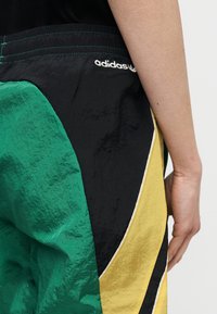 Κοντινό πλάνο ενός ατόμου που φοράει σορτς Adidas σε πράσινο, μαύρο και κίτρινο με λευκό λογότυπο, εμφανίζοντας μέρος του χεριού και του βραχίονα ενάντια σε απλό φόντο.