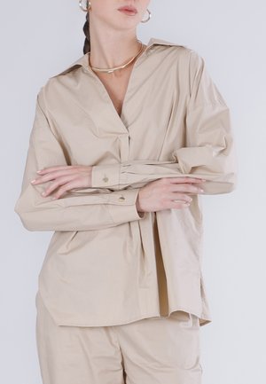 Nainen pukeutuneena beigeen väljään paitaan ja housuihin, kullanväriset renkorenkaat korvissa ja kaulakorussa, kädet ristissä vaaleaa taustaa vasten.