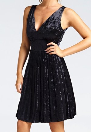 Kvinde iført ærmeløs sort fløjlsærklædning med dyb V-hals og plisseret nederdel, poserer med den ene hånd på hoften.