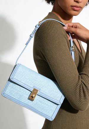 Frau trägt ein olivgrünes, geripptes Oberteil und hält eine hellblaue, strukturierte Handtasche mit goldener Schnalle über der Schulter vor weißem Hintergrund.