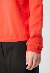 Main reposant sur le côté portant un pull en tricot à manches longues orange vif et un pantalon noir sur un fond clair uni.