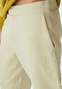 Pantalones color beige claro con una textura suave, que cuentan con un bolsillo lateral y un sutil logo en relieve "Calvin Klein" en la pierna izquierda.