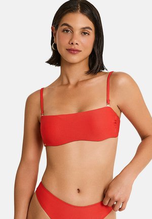 Jeune femme aux cheveux courts et foncés portant un haut et un bas de bikini rouge côtelé sur un fond clair uni.