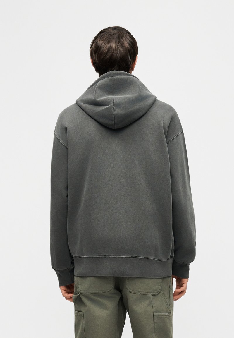 Person wearing a dark gray hooded sweatshirt and green cargo pants, standing facing away against a plain light background.