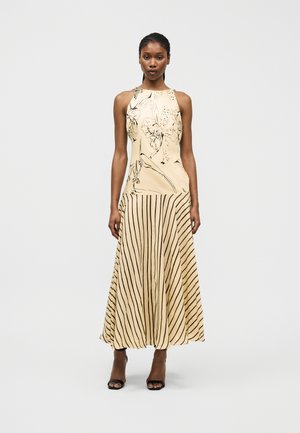 Žena nosi bezrukávové béžové šaty s kvetinovým vrškom a sukňou s vertikálnymi pruhmi, doplnené čiernymi sandálmi na vysokom podpätku.