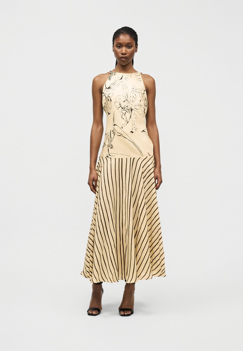 Γυναίκα που φοράει μπεζ αμάνικο φόρεμα με τοπ με floral σχέδιο και φούστα με κάθετες ρίγες, συνδυασμένο με μαύρες ψηλοτάκουνες σανδάλια.