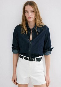 Cămașă din denim cu nasturi și mâneci suflecate, material bleumarin, cusături albastre deschise; asortată cu pantaloni scurți albi și o curea neagră.