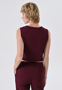 Top senza maniche bordeaux con scollo rotondo e design cropped; texture liscia, stile minimalista, abbinato a pantaloni coordinati.