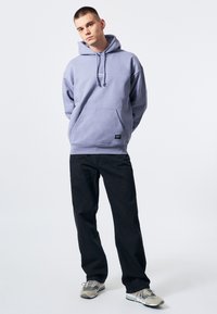Ljust lila hoodie med känguruficka, dragsko-huva och logodetalj, ihop med svarta byxor och grå sneakers.