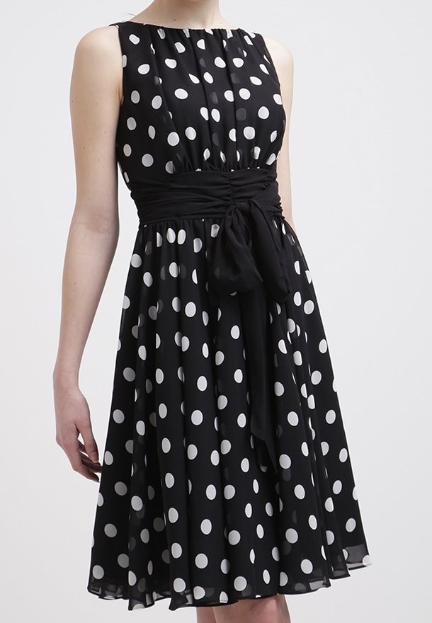 Robe noire sans manches à pois blancs, taille cintrée avec large ceinture noire nouée en nœud, jupe longueur genou.