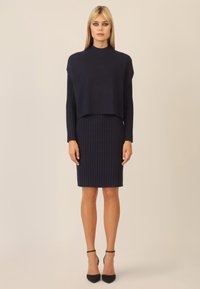 Marineblaues Strickkleid mit einem cropped Oberteil und geripptem Muster, ausgestattet mit langen Ärmeln und einem taillierten Rock, kombiniert mit schwarzen High Heels.