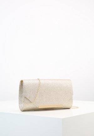 Guldglitter clutch taske med en tynd guldkæde-rem, der hviler på en hvid overflade mod en ensfarvet baggrund.