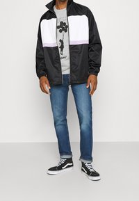 Schwarze Jacke mit weißen und lavendelfarbenen Streifen, graues Graphic-T-Shirt, blaue Jeans und schwarze High-Top-Sneaker mit weißen Akzenten.