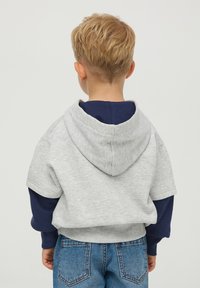 Szara bluza z kapturem i krótkimi rękawami, nałożona na granatową koszulę z długim rękawem, w połączeniu z niebieskimi dżinsami. Casualowy, luźny krój.