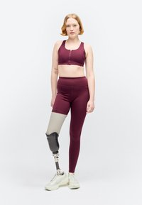 Femme en tenue de sport bordeaux debout avec une prothèse à la jambe gauche, chaussée de baskets blanches sur fond blanc uni.