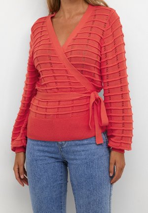 Koralfarvet strikket omslagsweater med teksturerede vandrette striber og sidelukning, båret med lyseblå jeans med høj talje på en stående person.