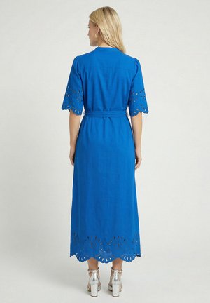 Femme aux longs cheveux blonds portant une robe midi bleu cobalt avec des manches et un ourlet festonnés à œillets, ainsi que des sandales à talons hauts argentées, de dos.