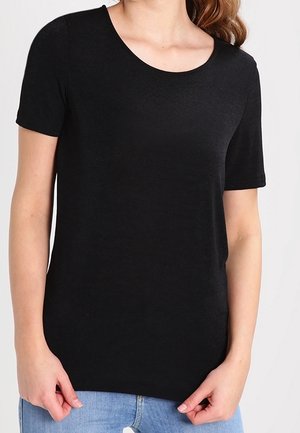 Kvinde iført en enkel sort kortærmet top og blå jeans, hænderne rører let ved kanten af toppen.
