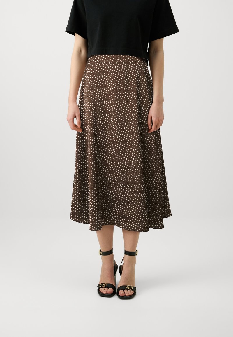 Femme portant une jupe midi marron à pois blancs, un top court noir et des talons noirs à bride cheville, debout devant un fond uni.