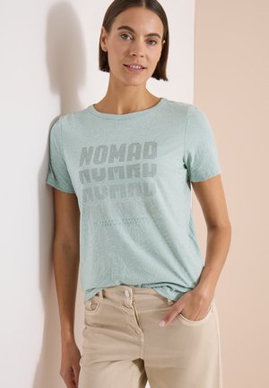Femme aux cheveux courts et bruns portant un t-shirt bleu clair imprimé "Nomad" et un pantalon beige, debout contre un mur blanc et beige.