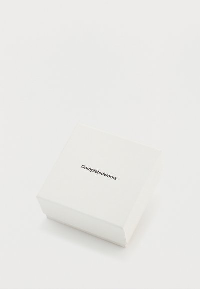 Cutie pătrată albă cu o textură netedă, având imprimat "Completedworks" cu un font negru îngroșat, poziționat ușor decalat față de centru.