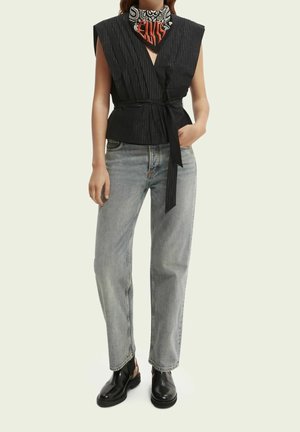 Femme portant un haut cache-cœur sans manches noir avec ceinture, jean gris clair, bottes noires et foulard noir et blanc à motifs avec texte rouge.