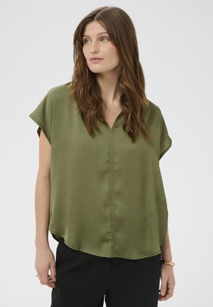 Pikkade brünettvärviliste juustega naine, kes kannab oliivrohelist lühikeste varrukatega pluusi ja musti pükse, ning vaatab veidi kõrvale.