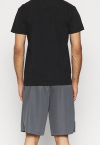 Mand iført en sort kortærmet skjorte og grå knælange shorts, stående med ryggen til mod en ensfarvet lys baggrund.