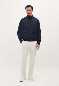 Marineblauwe bomberjack met een hoge kraag, twee borstzakken en een ritssluiting aan de voorkant. Combinatie met witte broek en canvas sneakers.