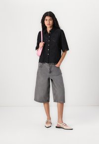 Svart bluse med eyelet-design og korte ermer, grå vide denimshorts, og beige leppestifter med stropp. Tilbehør inkluderer en liten rosa håndveske.