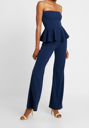 Femme portant un top bustier bleu marine à basque et un pantalon large assorti avec des sandales à talons hauts jaunes, debout sur un fond blanc.