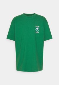 Camiseta de algodón verde con un corte holgado, que presenta un cuello redondo, mangas cortas y un estampado gráfico en el área del lado izquierdo del pecho.