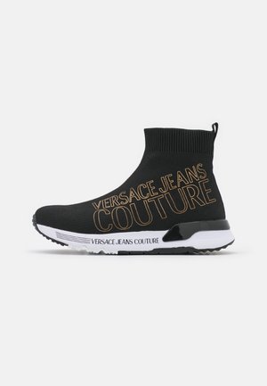 Czarny sneaker w stylu skarpetkowym z białą podeszwą, złoty napis „Versace Jeans Couture” z boku i czarna pętelka na pięcie.