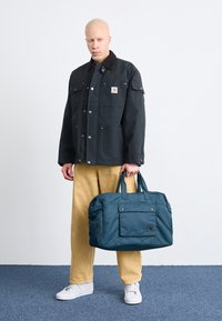 Jachetă neagră din pânză cu guler maro, două buzunare pe piept; pantaloni din catifea mustar; geantă duffel teal cu buzunar frontal; adidași albi.