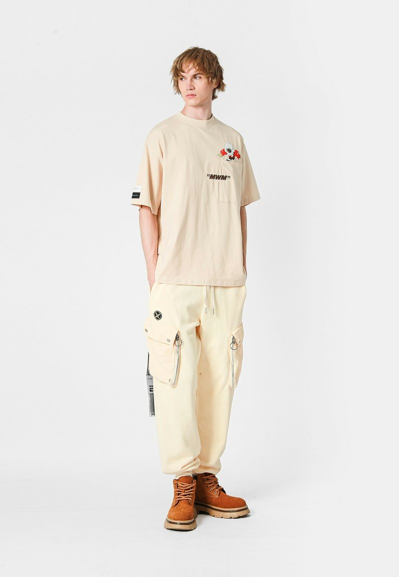 Camiseta oversize beige con gráfico floral y bolsillo, combinada con pantalones cargo crema con cremalleras y detalles negros, terminando con botas marrones.