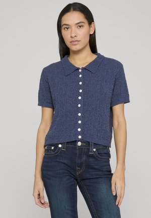Marineblaues, strukturiertes Kurzarmhemd mit einem Kragen und weißen Knöpfen vorne. Getragen mit dunklen Jeans.