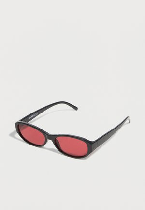 Čierne oválne slnečné okuliare s tenkými ramenami a červenými tónovanými šošovkami ležia na bielom povrchu.
